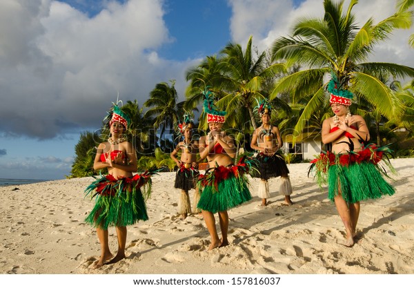 Polynesian\
Pacific Islanders Tahitian people dancing group wearing traditional\
colorful costumes  dancing on sand of a pacific island tropical\
beach with palm trees in the\
background.