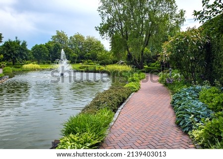A pond filled with water with a spray jet fountain in a park with pedestrian sidewalks made of stone tiles among different plants, landscape design of flower beds and trees.