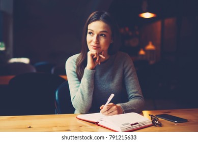 Pondering Attractive Young Woman With Pen In Hand Thinking On Plans And Writing List To Do In Notepad Enjoying Recreation Time In Stylish Coworking Space.Thoughtful Student Noting Text Information