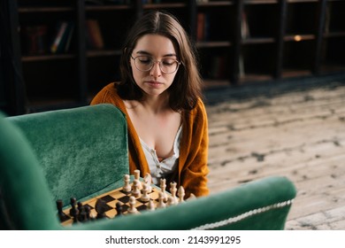 Pondering Attractive Young Woman Wearing Elegant Eyeglasses Thinking About Chess Move While Sitting On Wooden Floor, By Armchair In Dark Room Playing Logical Board Game Alone.