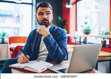 Pondering Bearded Creative Man In Denim Apparel Thinking On Productive Ideas For Developing Own Startup Project Sitting At Modern Laptop Computer And Notepad For Notes In Stylish Coworking Space