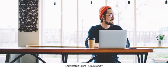 Pondering hipster guy thoughtful looking away and thinking about information database on laptop computer during distance job in coffee shop, front view of caucasian freelancer sitting indoors
