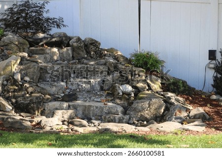 A pondless backyard waterfall. Three bronze turtle statues sit on the rocks and is surrounded with red mulch, juniper bushes, holly bushes, and a Japanese maple tree.