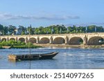 The Pont Wilson bridge spans the Loire River in Tours, France, showcasing its elegant stone arches and historic architecture. In the foreground, a traditional wooden boat floats on the river.