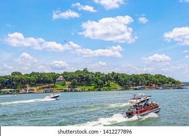 Pontoon Boat Full Of People And Two Speedboats Race Down Lake With Luxury Homes And Docks On Shore Under Bright Blue Sky With Clouds And Helicopter Above