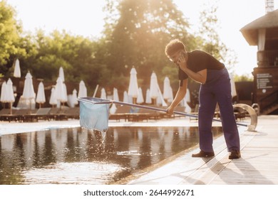Pool cleaner is scooping leaves from the pool surface and talking on the phone at the same time in a luxury resort
