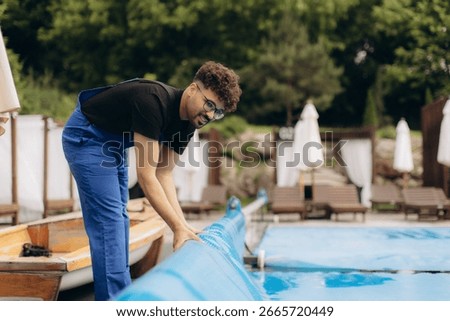 Pool maintenance worker rolling up a pool cover at a resort, ensuring cleanliness and safety for guests enjoying summer leisure by the blue water