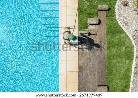 A pool maintenance worker uses a net to clean the surface of a clear blue swimming pool, surrounded by green grass and stone borders on a bright day.