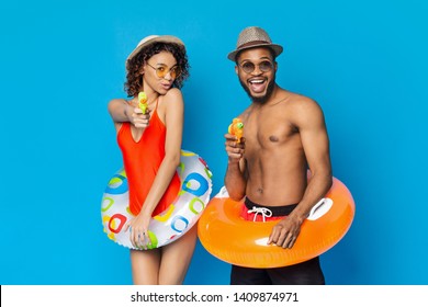 Pool Party Fun. Black Couple With Inflatable Rings Shooting At Camera With Water Guns, Blue Studio Background