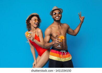 Pool party time. Happy millennial couple in swimwear drinking summer cocktails, blue studio background
