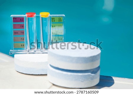 Pool water test kit with ph and chlorine color chart beside white tablets on pool edge. Blue water in background