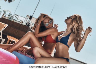 Poolside fun. Two beautiful young women in bikini smiling while sitting by the pool outdoors