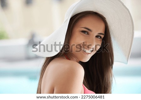 Poolside is the only place to be. Portrait of a smiling young woman sitting by a pool wearing a sunhat.