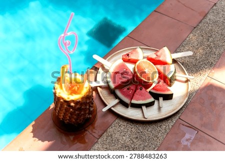 Poolside scene with a pineapple drink and a platter of watermelon and orange slices on sticks, set on a sunlit tiled surface.