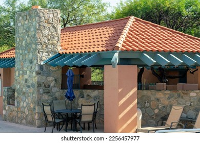 Poolside villa or kitchen outside in a public community area with small rock chimney and adobe roof tiles and stucco. Recreation area in a park or waterway with tables and chairs visible.