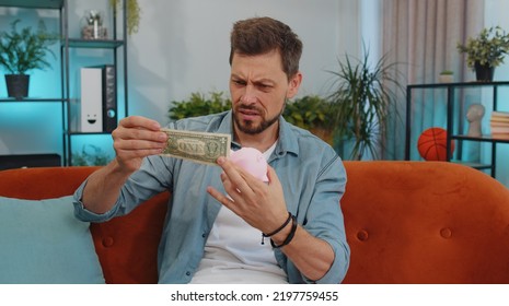 Poor Adult Man Insufficient Amount Of Money, Holding Piggybank And One Dollar Banknote At Home. Financial Crisis. Bankruptcy. Poverty And Destitution. Young Guy At Home Apartment Living Room On Couch