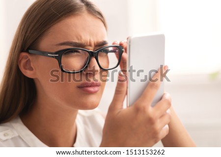 Poor Eyesight Concept. Girl Looking At Cellphone Screen Through Eyeglasses Having Vision Problem Indoor. Selective Focus