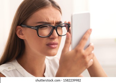 Poor Eyesight. Girl Squinting Eyes Looking At Smartphone Screen Through Eyeglasses Sitting Indoor. Selective Focus