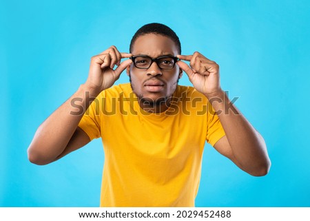 Poor Eyesight Problem. Black Guy In Glasses Squinting Eyes Looking At Camera Posing In Studio Over Blue Background. Vision Health Issue Concept. Portrait Of Nearsighted Man Wearing Eyeglasses