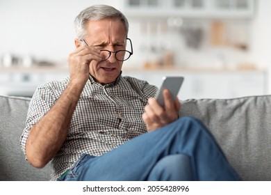 Poor Eyesight. Senior Man Squinting Eyes Reading Message On Phone Wearing Eyeglasses Having Problems With Vision Sitting On Couch At Home. Ophtalmic Issue, Bad Sight In Older Age Concept - Shutterstock ID 2055428996