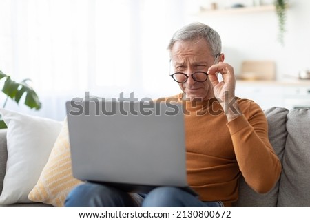 Poor Eyesight. Senior Man Squinting Eyes Using Laptop Wearing Eyeglasses Having Problems With Vision Sitting On Couch. Ophtalmic Issue, Bad Sight In Older Age, Macular Degeneration Concept