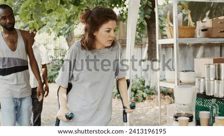 Poor, hungry Caucasian lady on crutches approaches an outdoor food bank to receive free food assisted by black woman. Volunteers serving meals and nourishments to underprivileged and homeless people.
