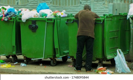Poor and hungry homeless man in dirty clothes looking for food in the dumpster on the urban street in the city. Social problems of modern society. Lifestyle of tramp, living in the streets. - Shutterstock ID 1748095844