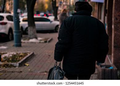 Poor Pensioner In Big Black Jacket With Black Bag Going On The Street. Back View