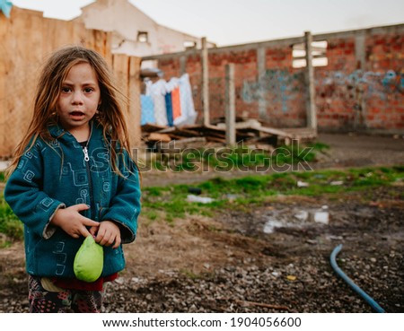 A poor and sad gypsy little girl is standing in a Roma settlement, holding a toy in her hand and looking at the camera