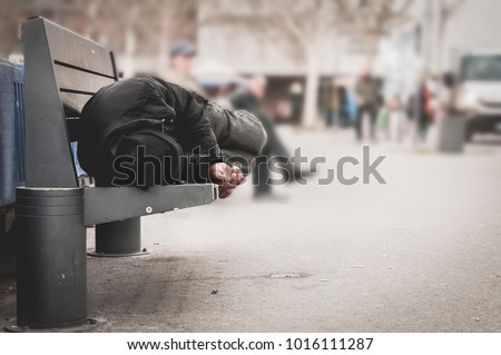 Poor tired depressed hungry homeless man or refugee sleeping on the wooden bench on the urban street in the city, social documentary concept