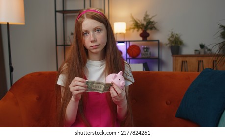 Poor Young Teen Child Girl Insufficient Amount Of Money, Holding Piggybank And One Dollar Banknote At Home. Financial Crisis. Bankruptcy. Poverty And Destitution. Ginger Kid In Living Room On Couch