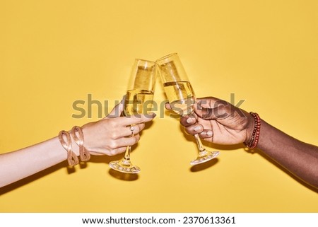Pop color closeup of two hands clinking tall champagne glasses against vibrant yellow background