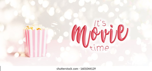 Popcorn Bag With White And Pink Stripes At The Festive Lights Bokeh Backdrop. Romantic Movie Night And Snack Concept With It's Movie Time Wording. Banner Wide Screen Format