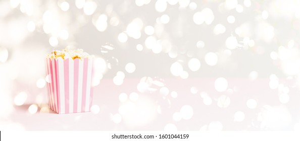 Popcorn Bag With White And Pink Stripes At The Festive Lights Bokeh Backdrop. Empty Minimalistic Background. Romantic Movie Night And Snack Concept. Banner Wide Screen Format