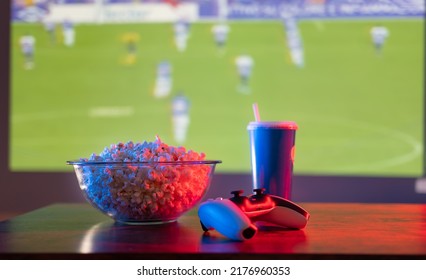 Popcorn In A Glass Bowl, A Carbonated Drink In A Plastic Glass, A Gamepad On The Table Against The Background Of A Large TV With A Football Match. Sports, Football, Video Games, Fast Food.