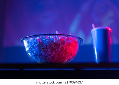 Popcorn In A Glass Bowl And A Drink In A Plastic Cup On The Table. Neon Lighting. Cozy Watching Your Favorite Movies And TV Shows With Fast Food. Rest, Comfort, Day Off.