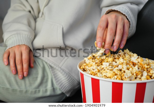 Popcorn paper bucket in the hands of a young girl\
preparing to watch a movie. Showtime. Eating delicious unhealthy\
sweet snacks. Going to cinema for a new film. Rest and\
entertainment. Popcorn\
closeup