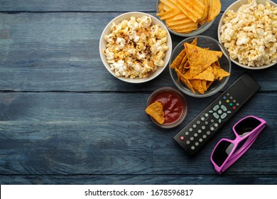 Popcorn, Various Snacks, 3D Glasses And A TV Remote Control On A Blue Wooden Table. Concept Of Watching A Movie Or Video At Home. Top View