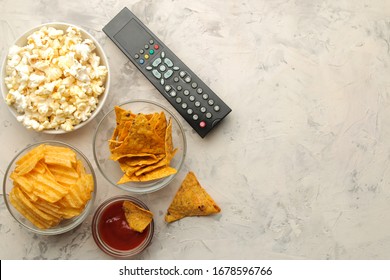 Popcorn And Various Snacks And A Tv Remote Control On A Light Concrete Table. Concept Of Watching A Movie Or Video At Home. Top View