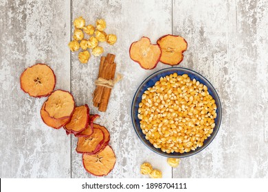 Popped and unpopped corn kernals with apple slices, cinnamon, unpopped corn kernals on white wooden background, shot from above