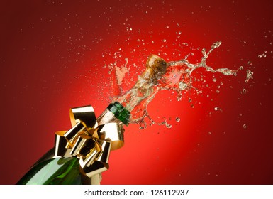 Popping Cork From Champaign Bottle With Gold Bow On It And Splashes All Around The Red Background