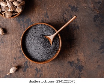 Poppy seeds in a wooden bowl. Black, dark gray small round seeds of dried poppy seed pops. Close up view. Brown wooden background, tea spoon. Cooking, baking ingredient. Organic food. Copy space.