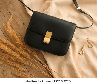 Popular Black Bag Made Of Genuine Leather With Gold-colored Fittings, And Decor In The Form Of Earrings Accessories, Beige Background Decorated With A Reed Stem.