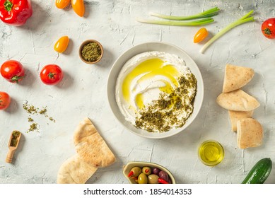 Popular middle eastern appetizer labneh or labaneh, soft white goat milk cheese with olive oil, hyssop or zaatar, served with many fresh vegetables on a wooden plate over grey table, flaylay.
						