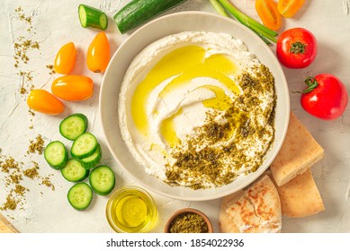 Popular middle eastern appetizer labneh or labaneh, soft white goat milk cheese with olive oil, hyssop or zaatar, served with fresh vegetables on a wooden plate over grey table, flaylay. Close up
					