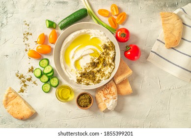 Popular middle eastern appetizer labneh or labaneh, soft white goat milk cheese with olive oil, hyssop or zaatar, served with many fresh vegetables on a wooden plate over grey table, flaylay.
