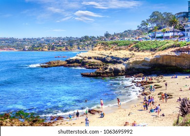 Popular seaside town of La Jolla Cove in San Diego with a crowd on the beach, canoers on the waters, and a group of sea lions resting on the rocks.