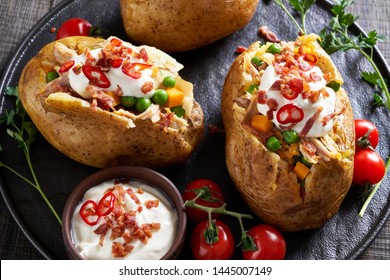 Popular street food: twice baked russet potatoes with bacon, cheddar, sour cream, chicken and green peas, served on a black plate sprinkled with roasted bacon, hot chili, with cherry tomatoes  - Shutterstock ID 1445007149