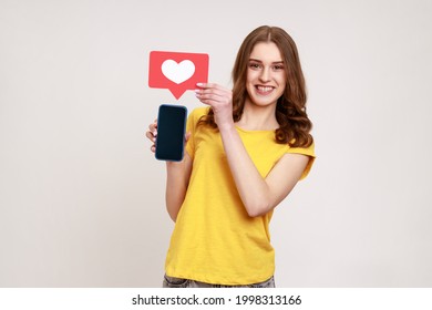 Popularidad en medios sociales. Mujer sonriente de cabello ondulado en camiseta sosteniendo el corazón de la red Como icono y mostrando el teléfono inteligente con pantalla en blanco. Estudio cubierto aislado en fondo gris.