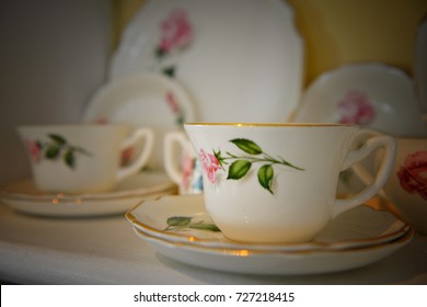 Porcelain cups and saucers stacked on a shelf with a yellow wall in the background. The delicate old fashioned bone china dishes have a red rose and green leaf pattern with gold trim on the edges. - Powered by Shutterstock